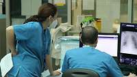 Ein Arzt in Krankenhauskleidung sitzt vor Monitoren. Auf einem ist ein Röntgenbild zu sehen. Links daneben steht eine Ärztin. 