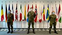 Three service members with attached Cross of Honour of the Bundeswehr are standing in front of different national flags