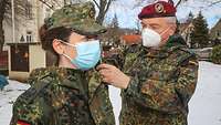 Eine Soldatin mit Mundschutz bekommt von einem Soldaten mit Mundschutz die Schulterstücke ausgetauscht.