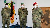 Drei Generale mit Mund-Nasenschutz stehen nebeneinander vor der Truppenfahne und einem aus Holz geschnitzten Divisionswappen.