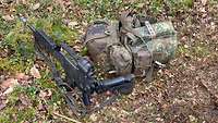 Auf Waldboden: Ein aufgestelltes Gewehr, daneben in Flecktarn verpackt Ausrüstungsgegenstände.
