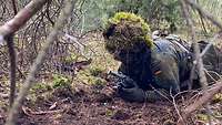 Ein Soldat mit getarntem Gefechtshelm und Gewehr robbt über Waldboden.