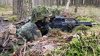 Ein getarnter Soldat liegt mit seinem Gewehr im Anschlag im Wald in einer Stellung.