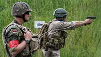 Zwei Soldaten mit Helm und Ausrüstung stehen auf einer Wiese. Der rechte hält eine Pistole im Anschlag und feuert.