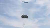 Am Himmel schwebt ein Mensch an einem Fallschirm, im Hintergrund ein großes Propellerflugzeug