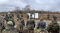 Zahlreiche Soldaten aus verschiedenen Ländern stehen in der Heide und schauen auf Tafeln, neben denen andere Soldaten stehen.