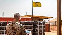 Ein Soldat steht mit dem Rücken zur Kamera und hält eine Postkarte in der Hand. Im Hintergrund die Feldpoststelle