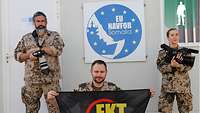 Zwei Soldaten und eine Soldatin stehen vor dem EU-NAVFOR-Wappen. Einer der drei kniet mit der EKT-Flagge in der Mitte 