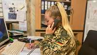 Eine Soldatin sitzt an einem Schreibtisch, hält einen Telefonhörer in der Hand und notiert sich etwas in ein Buch