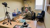 Ein Soldat sitzt am Schreibtisch vor einem Laptop. Auf dem Tisch neben ihm steht eine Kamera auf einem Stativ