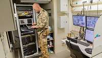 Ein Soldat steht an einem Laptop neben einer Serveranlage. Hinter ihm stehen drei Bildschirme auf einem Schreibtisch