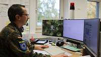 : Ein Soldat sitzt an einem Schreibtisch. Vor ihm sind drei Monitore zu sehen