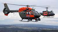 In der Luft: zwei in Flecktarn lackierte Hubschrauber mit jeweils einer orangefarbenen Fläche, darauf die Buchstaben SAR.