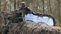 Ein Geländewagen mit Tarnnetzen steht im Wald. Aus dem offenen Dach schaut ein Soldat mit einem Gewehr im Anschlag.