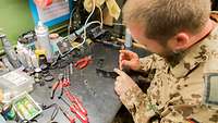 Ein Soldat sitzt an einer Werkbank und repariert eine Sonnenbrille. Auf dem Tisch liegen Utensilien für die Reparatur