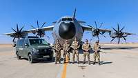 Vor einem Transportflugzeug stehen fünf Soldatinnen und Soldaten im Halbkreis. Neben ihnen steht ein grünes Militärfahrzeug 