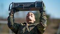 Ein Soldat stemmt eine metallene Kiste mit beiden Armen über seinem Kopf und presst dabei die Lippen zusammen.