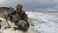 A soldier in blue undress uniform, camouflage flak jacket, and combat helmet stands besides a small boat on a beach.