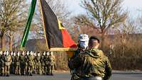 Zwei Soldaten überreichen sich eine Fahne auf einem Platz. Im Hintergrund ist eine Formation Soldaten angetreten.