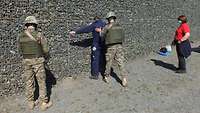 Zwei Soldaten kontrollieren einen Mann, der mit erhobenen Armen an einer Wand steht. Eine Frau mit roten Shirt steht daneben.