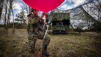 Ein Soldat kniet auf einer Wiese und füllt einen großen, roten Wetterballon mit einem Gasfüllschlauch.