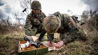 Zwei Soldaten knien auf einer Wiese und übertragen Messewerte aus Unterlagen.