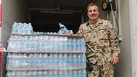 Ein Soldat mit einer Wasserflasche in der Hand steht neben einer Palette mit Wasserflaschen und lächelt in die Kamera
