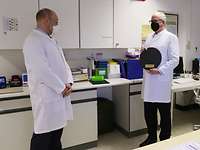 Zwei Männer stehen in einem Labor. Der Rechte hält eine kreisrunde schwarze Schieferplatte in der Hand.