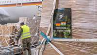 Ein Soldat und mehrere Kartons vor einem Airbus A350, im Vordergrund eine Postkarte mit dem Text „#Hilfseinsatz“ an einem Karton