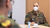 Ein Soldat sitzt am Tisch und schaut in Richtung eines Generals, vor ihm steht eine Kaffeetasse