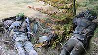 Zwei Soldaten liegen an einem Waldrand auf einer Anhöhe und beobachten mit Ferngläsern eine Straße.