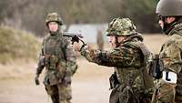 Ein Soldat schießt im Stehen mit einer Pistole auf dem Truppenübungsplatz, zwei Soldaten stehen daneben und beobachten ihn