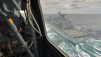 Aus dem Seitenfenster des Hubschraubers heraus ist ein graues Schiff im Vorbeiflug zu sehen.