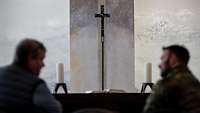 Altar mit zwei Kerzen und einem Kreuz aus Metall. Davor sitzen ein Zivilist und ein Soldat.