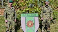 Zwei Soldaten in Uniform stehen draußen neben einem Wappen auf der grünen Wiese. 