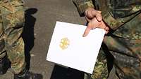 Ein Soldat hält eine Urkunde mit dem Bundeswappen auf dem Deckel hinter dem Rücken in der Hand.