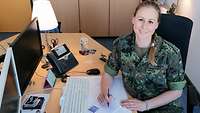 Eine Soldatin sitzt an einem Schreibtisch und lächelt in die Kamera