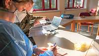 Eine Person in medizinischer Schutzkleidung sitzt an einem Tisch und macht handschriftliche Notizen.