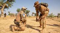 Ein malischer Soldat beugt sich in der Wüste zu einem knienden deutschen Soldaten herunter und unterhält sich mit ihm