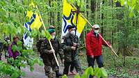 Zwei Männer in Uniform und einer in roter Jacke tragen Kreuz und Fahnen durch den Wald.