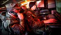 In einem Panzer sitzen bei rotem Licht zwei Soldaten vor einer Rechneranlage, einer mit Funkgerät.