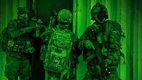 Drei Soldaten mit taktischer Ausrüstung, Waffen und Nachtsichtgeräten stehen an der Tür eines Gebäudes hintereinander.