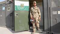 Ein Soldat mit einem Messgerät in der Hand kommt aus einem Container, an dessen Tür eine Sachsen-Flagge hängt 