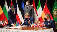 Ein Soldat umgeben von Flaggen verschiedener Nationen unterschreibt ein Dokument an einem Tisch im Beisein von zwei Soldaten