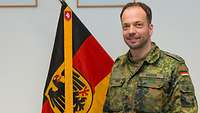 Ein Soldat steht vor der Deutschlandflagge und lächelt in die Kamera.