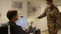 Ein Soldat überreicht einem Zivilisten, der am PC sitzt, eine Mappe. Beide tragen Mund-Nasen-Schutz