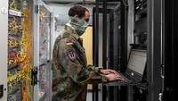 Ein Soldat in Uniform steht an einem Computer in einem Serverraum.