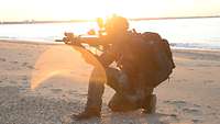 Ein Soldat in Uniform kniet mit seinem Gewehr am Strand und zielt.