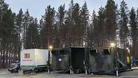 Two green containers and one white container in front of trees, a car standing next to them