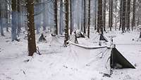 Mehrere Zwei-Mann-Zelte in einem Waldstück. Es liegt Schnee - auch auf den Zelten.